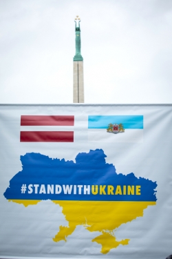 Plakāts ar Latvijas un Rīgas karogiem un uzrakstu #standwithukraine pie Brīvības pieminekļa